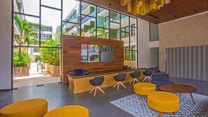 A modern reception area with a wooden reception desk, multiple seating options, and large windows offering views of greenery and buildings. - Premium Downtown PH +Private Pool +Gym+PKG+Rooftop (Playa del Carmen)