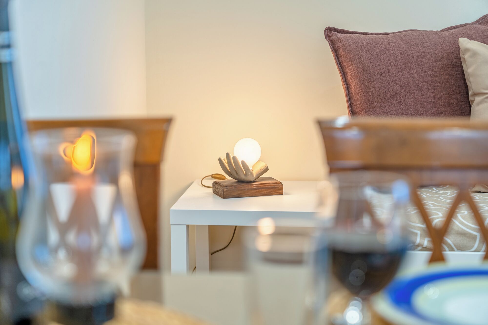 A cozy room with a bedside table holding a decorative object, a lamp, and a glass of wine.