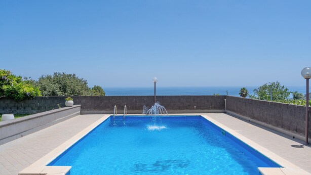 Une piscine rectangulaire avec une fontaine au centre, entourée d’un petit muret en pierre, offrant une vue dégagée sur l’océan en arrière-plan.