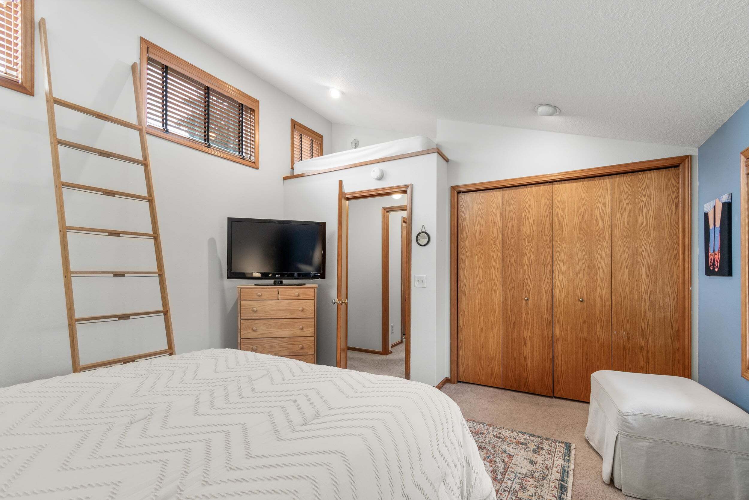 A bedroom with a bed, a TV, a wardrobe, a dresser, a ladder, and a door leading to another room.