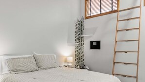 A bedroom with a bed, a bedside lamp, a ladder, a window with blinds, and a wall-mounted thermostat.