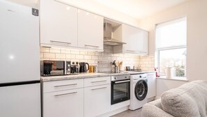 Een compacte keuken met witte kastjes, een inbouwoven, een wasmachine en een raam dat voor natuurlijk licht zorgt.