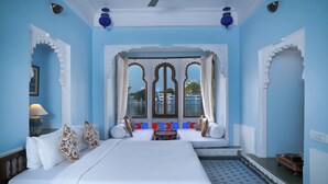 A room with a large bed, a small table, and a view through a window with decorative arches. - KANKARWA HAVELI (Udaipur)