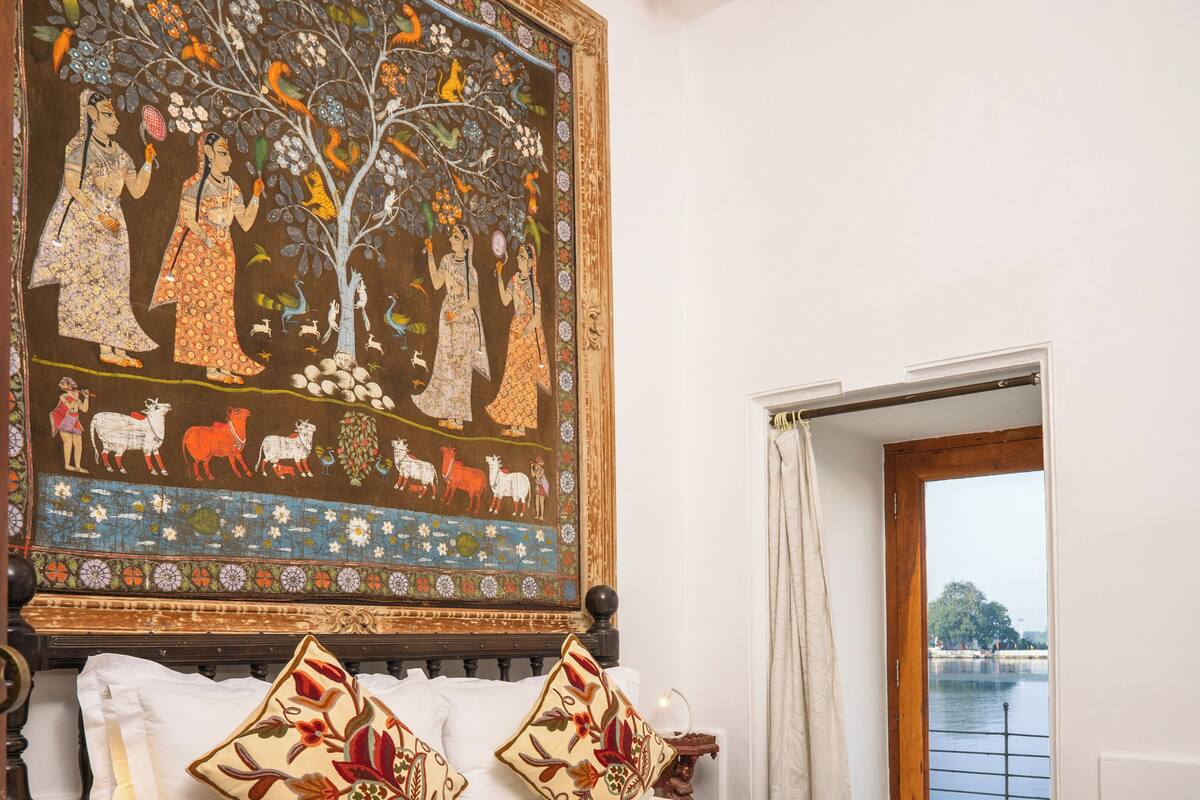 A bedroom with a large tapestry, a bed with white linens, and a view of a body of water through an open door.