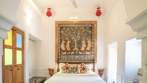 A bedroom with a bed, a ceiling fan, a decorative tapestry, and a wooden door. - KANKARWA HAVELI (Udaipur)