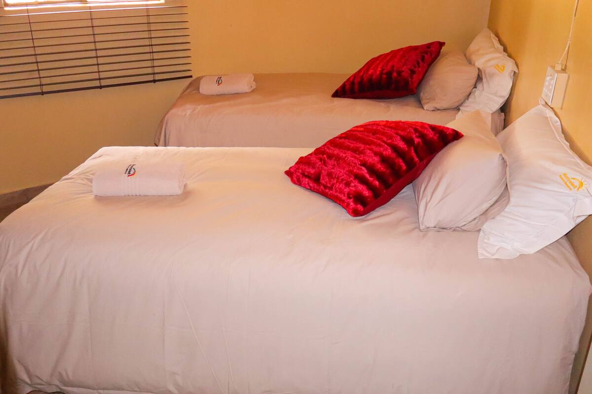 Two beds with white linens and red pillows, a window with blinds, and a wall socket.