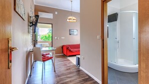 A hallway with a wooden floor, a dining area with a table and chairs, a red sofa, and a glass-enclosed shower area. - Apartment Pino by Interhome (Umag)