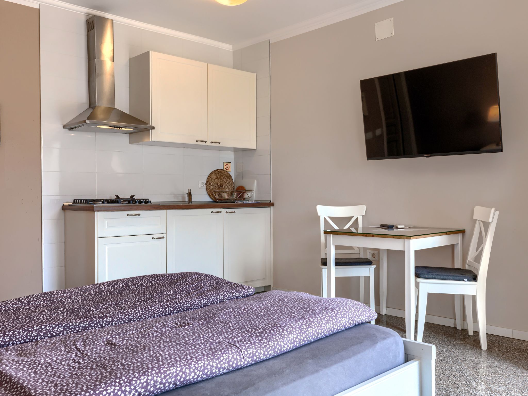 A compact room with a bed, a small table, two chairs, a TV, a kitchen area with cabinets and a stove.