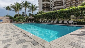 A rectangular swimming pool with clear blue water, surrounded by a paved deck with lounge chairs and a thatched-roof structure. - Experience coastal comfort at The Somerset, your oceanfront haven on Marco Island (Marco Island)