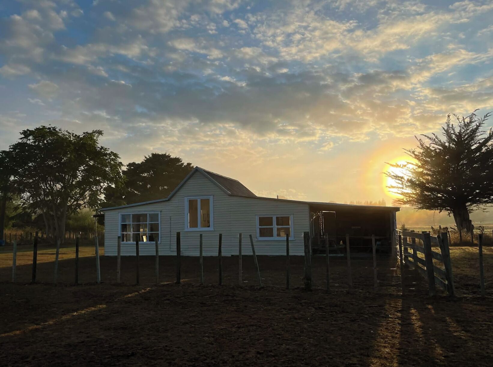 A single-story house with a gabled roof, surrounded by a wooden fence, set in a rural landscape with trees and a clear sky.