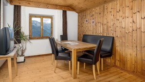 Ein Zimmer mit Holzwänden, einem Esstisch, Stühlen, einem Fernseher und einem Fenster mit Blick auf die Berge.