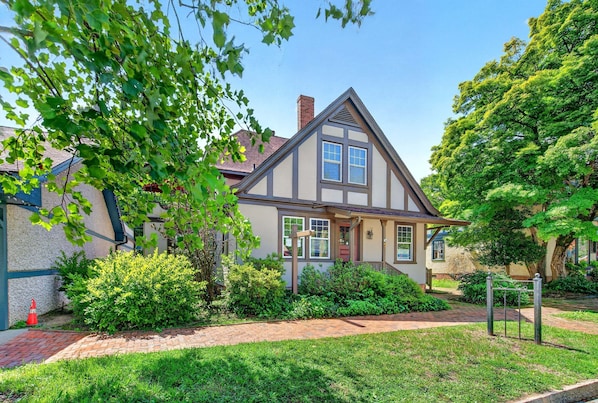 A two-story house with a gabled roof, a brick pathway leading to the entrance, and a well-maintained garden with various shrubs and a tree.