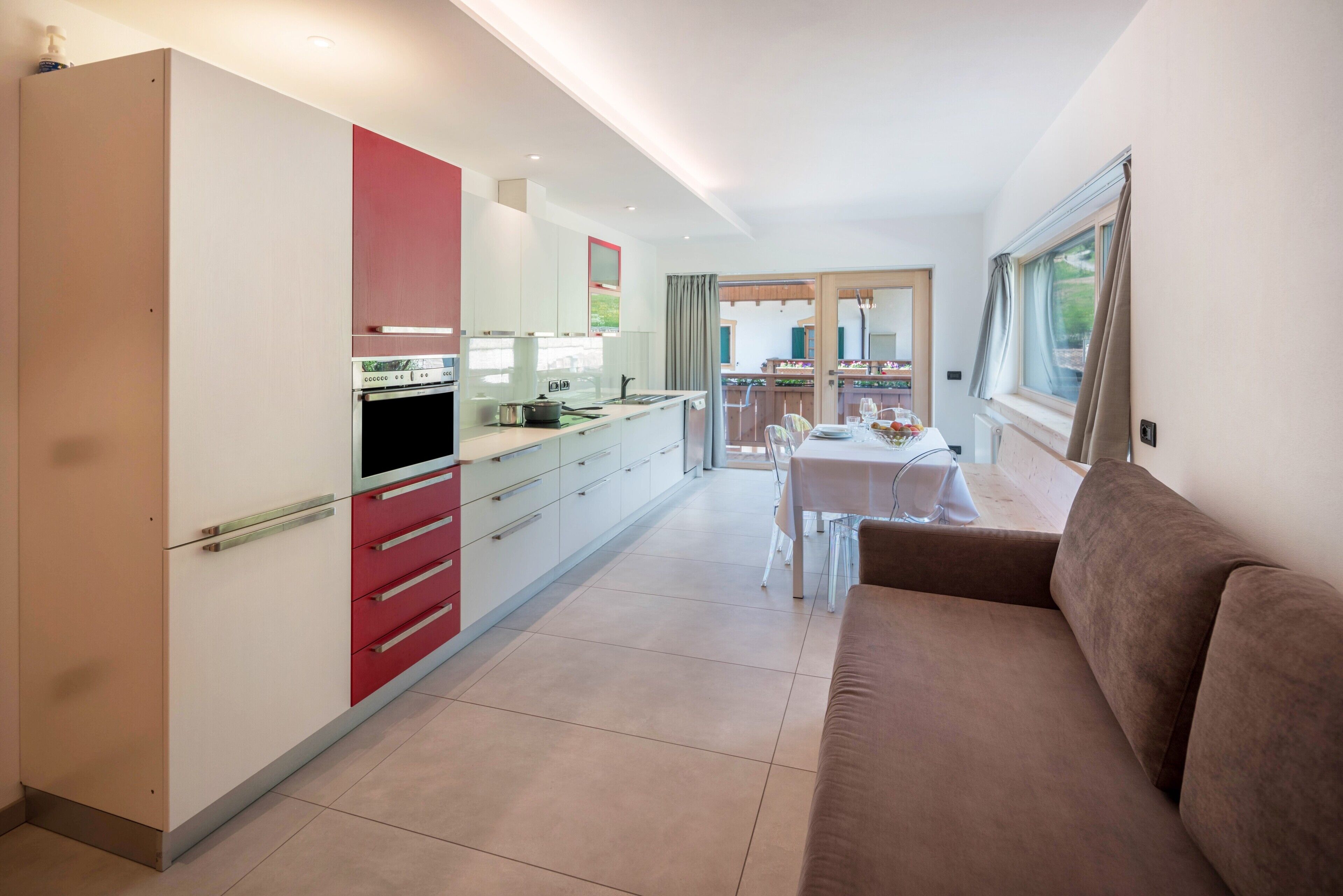 Une cuisine moderne avec des meubles blancs et rouges, un coin repas avec une table et des chaises, et un canapé marron.