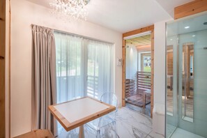 Una habitación moderna con una mesa de madera, una silla transparente y un ventanal que da a un balcón con terraza de madera.