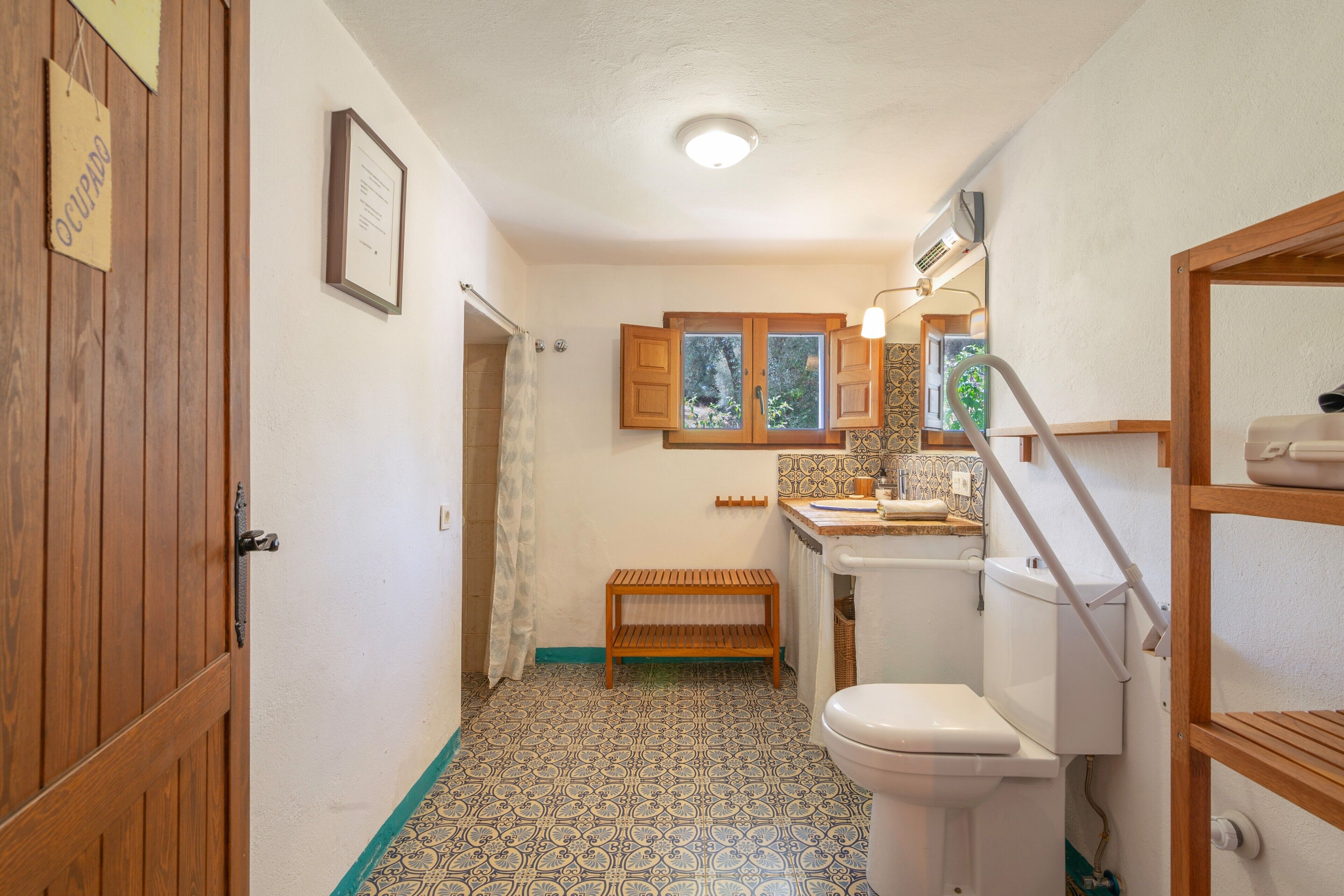 Un baño pequeño y funcional con inodoro, un banco de madera, ventana con vistas y puerta de madera.