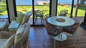 Amplio espacio interior con grandes ventanales con vista a un lago, amueblado con sillas de mimbre y una mesa redonda.