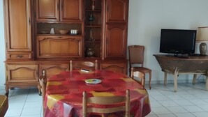 Un comedor con una mesa redonda cubierta con un mantel estampado, sillas de madera, un armario con estantes y un televisor sobre un mueble.