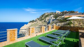 Eine Terrasse mit Liegestühlen, einer Steinmauer und Blick auf das Meer sowie einen Hügel mit Gebäuden.