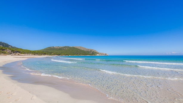 Ein Strand mit klarem Wasser, einem Sandufer und einem grünen Hügel im Hintergrund.