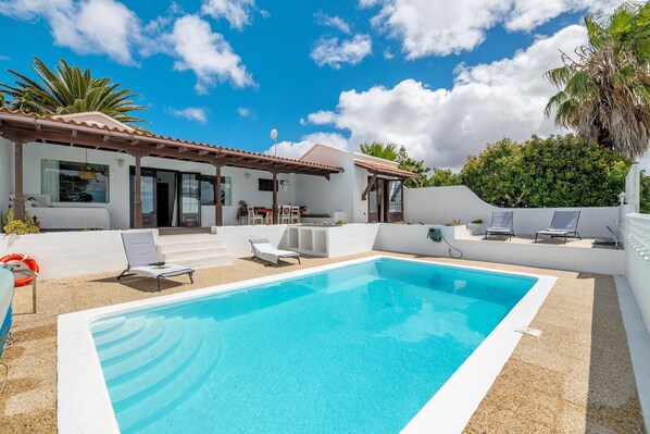 A swimming pool with lounge chairs, a covered patio, and a white building with large windows.