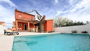 Piscina com espreguiçadeiras, um edifício com telhado de azulejos e uma árvore em primeiro plano.