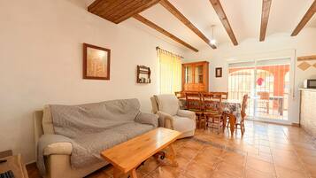 A living room with a sofa, armchair, wooden coffee table, and a dining area with chairs and a cabinet.