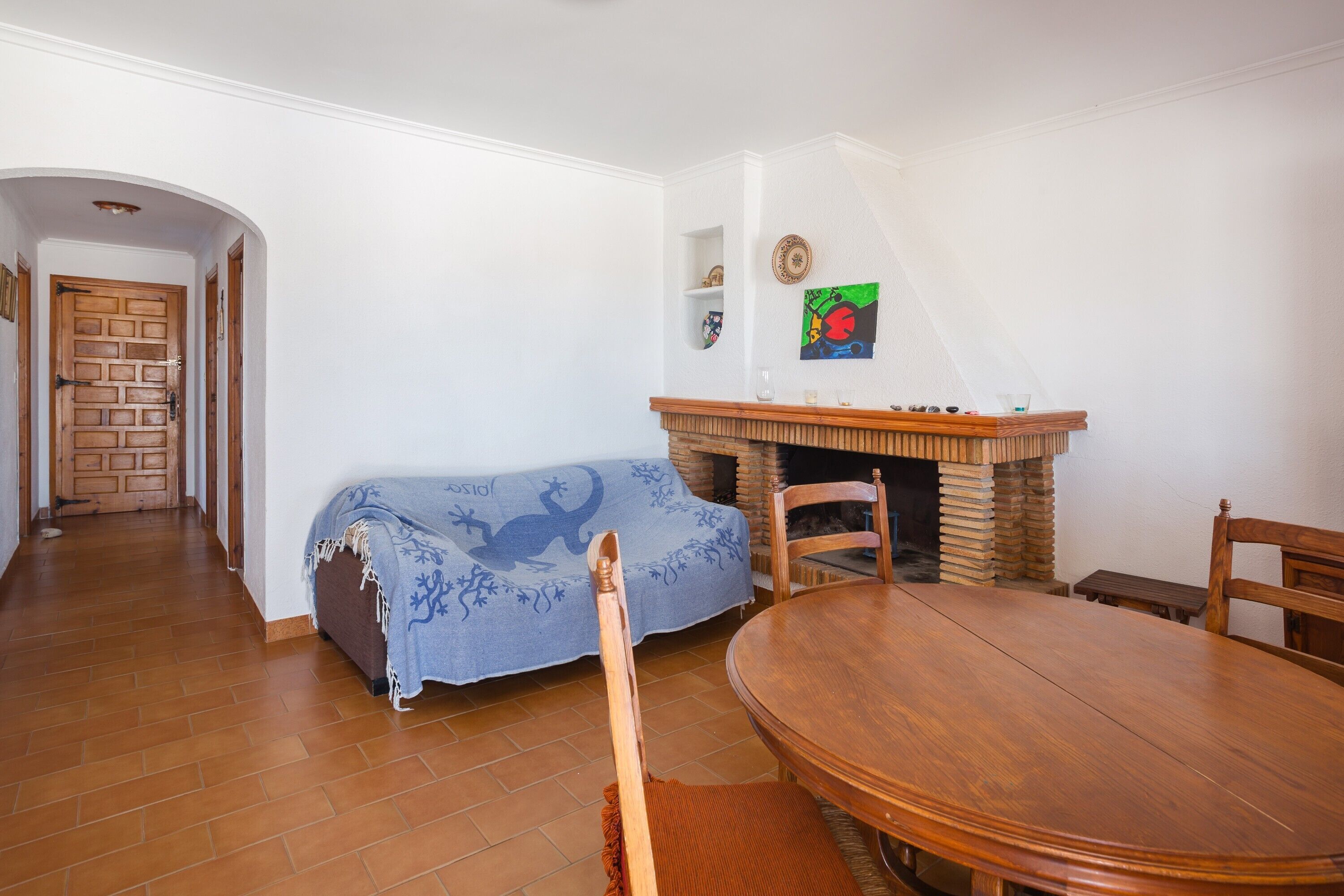 Ein Essbereich mit einem runden Holztisch und Stühlen, einem Kamin aus Backsteinen sowie einer blauen Couch mit Decke.