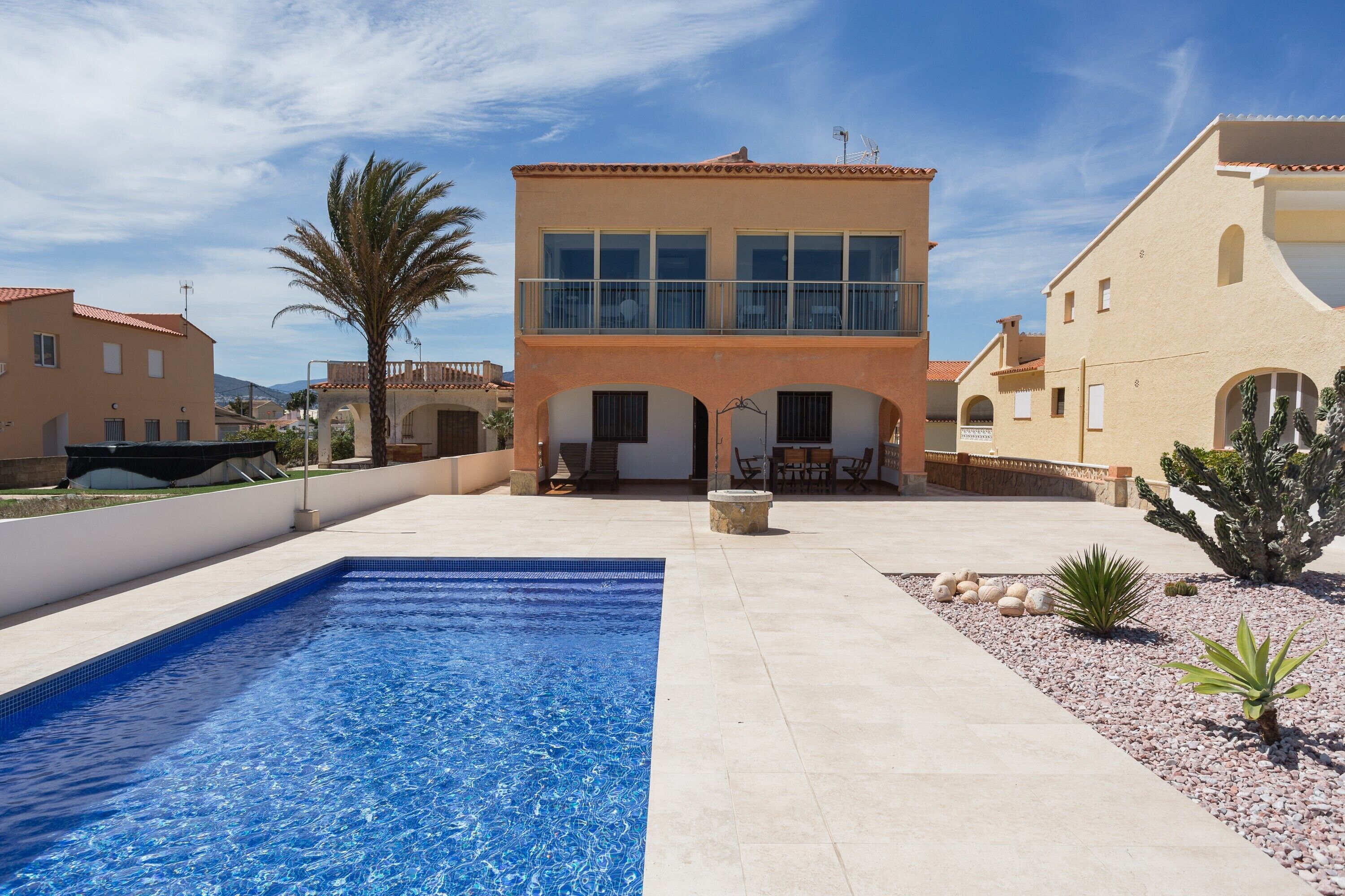 A swimming pool with a clear blue water surface, a two-story building with large windows and a balcony, and a desert garden with cacti and a palm tree.