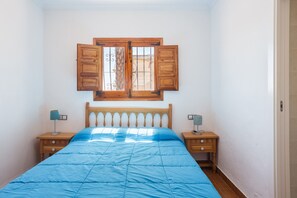 A bedroom with a bed, two bedside tables, a window with wooden shutters, and a wall-mounted light fixture.