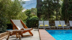 Un espace aménagé au bord de la piscine, avec une chaise en bois, un escalier métallique pour la piscine et des chaises longues blanches dotées de coussins jaunes.