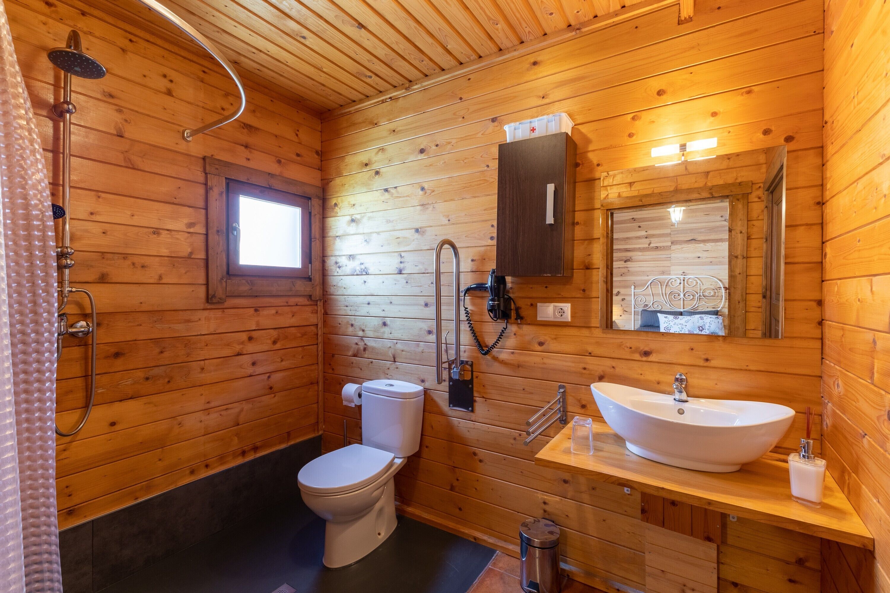 A wooden cabin bathroom with a toilet, sink, shower, and a window.