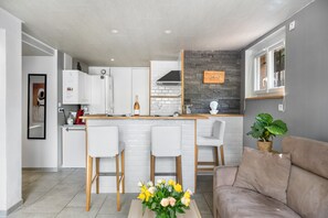 Una cocina moderna con armarios blancos, encimera de madera y salpicadero de piedra. Hay una zona de comedor con mesa y sillas blancas, y un sofá color beige en la sala de estar.