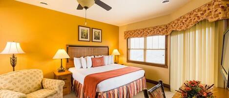 A hotel room with a bed, two bedside lamps, a ceiling fan, a window with blinds, a floral arrangement, and a wooden chair.