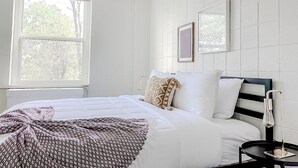 A neatly made bed with patterned bedding, a bedside table with a lamp, and a window with blinds. - Sleepover | Lavish 1BD/1BA + Gym Cincinnati (Cincinnati)