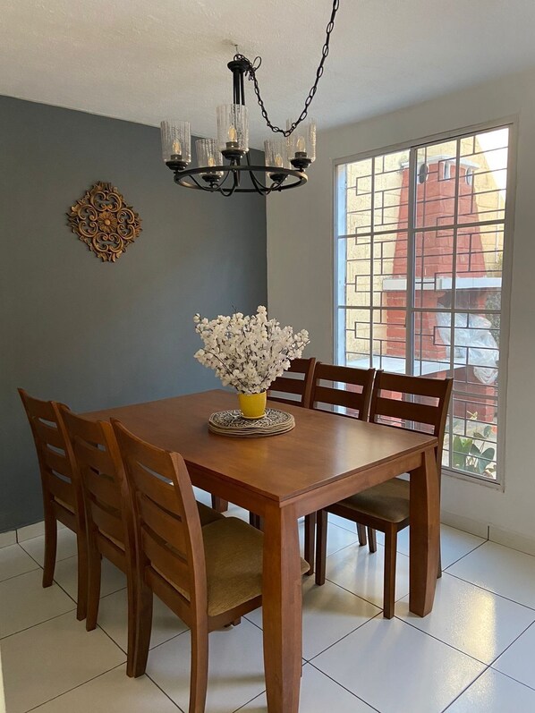 A dining room with a wooden table, six chairs, a yellow vase with white flowers, a decorative wall piece, and a window with a view of a building. - Charming 3-bedroom house in enchanting Santa Ana with AC, WiFi (Santa Ana)