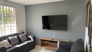 En stue med en grå sofa, et fjernsyn monteret på væggen, et træ-TV-bord og et vindue med metalgitter.