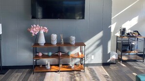 A modern living room with a flat-screen TV mounted on a grey brick wall, a wooden console table with decorative items, and a coffee machine on a cart. - Sleepover | Pristine 1BD/1BA + Gym - Cincinnati (Cincinnati)