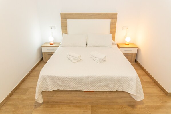 Un lit bien fait, avec une couette blanche et deux serviettes pliées, une tête de lit en bois et des lampes de chevet posées sur des tables de chevet en bois.