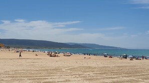 En bred sandstrand med mennesker og parasoller, en klar himmel og fjerne bjerge i horisonten.