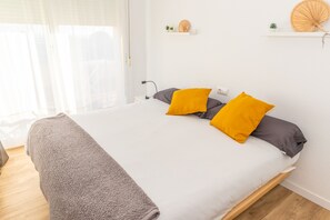 Una cama bien tendida con un edredón gris y dos almohadas, una amarilla y otra gris, en una habitación luminosa con una pared blanca y un pequeño estante con objetos decorativos.