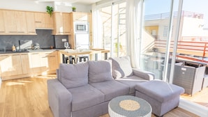 Ein modernes Wohnzimmer mit einem grauen Ecksofa, einem runden gewebten Couchtisch und einer Glastür zum Balkon.