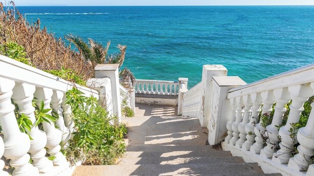 Eine Treppe führt zu einem Balkon mit Blick auf ein türkisblaues Meer.