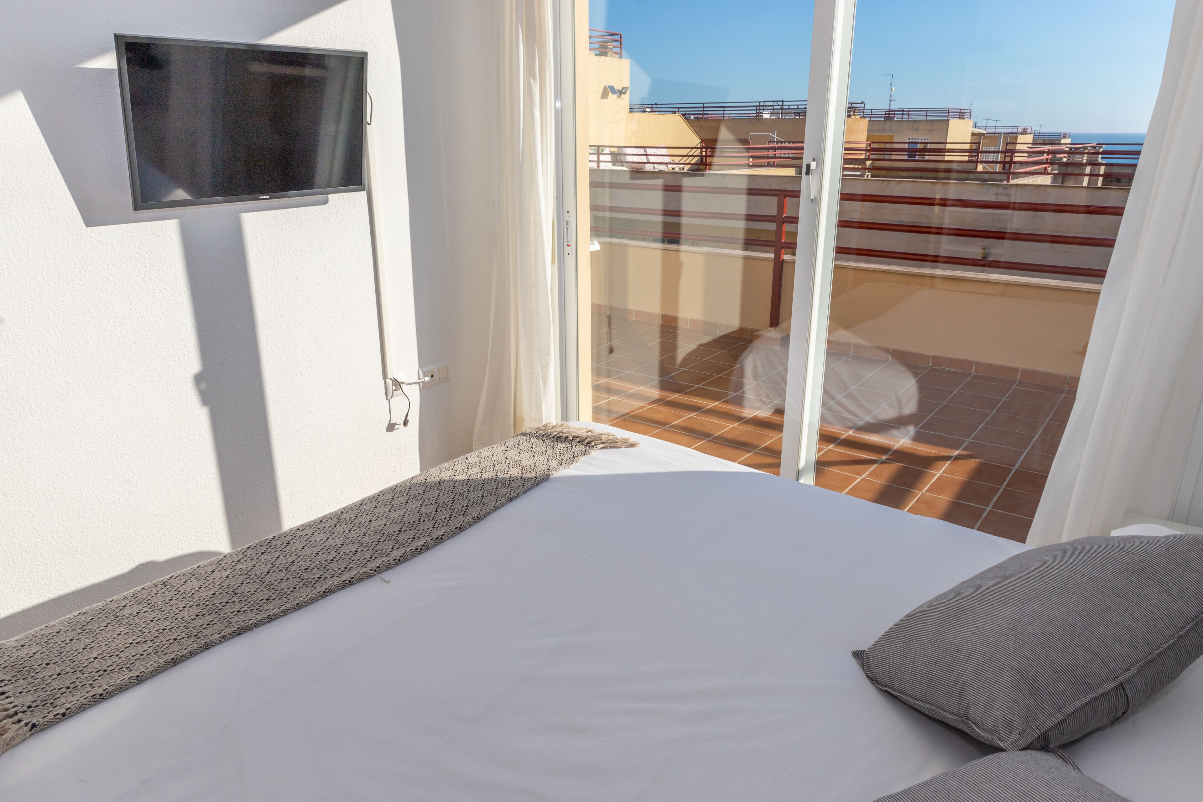 A hotel room with a bed, a TV mounted on the wall, and a balcony with a view of the sea.