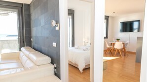 A modern hotel room with a balcony, a bed with white linens, a dining area with a table and chairs, and a TV mounted on the wall.