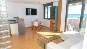 A modern living room with a glass coffee table, white sofa, and a dining area with a TV and a plant.