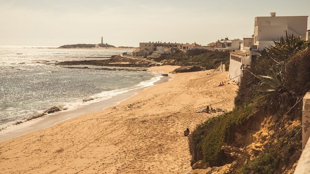 A coastal beach with buildings, a lighthouse, and rocky terrain. - Apartamento Sereno 5 | Piscina, Garaje, Terraza y Playa (Los Caños de Meca)