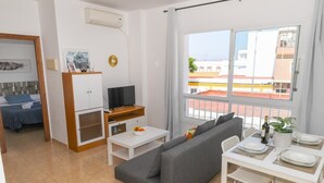 Een moderne woonkamer met een grijze bank, een televisie op een houten dressoir, een eettafel met stoelen en een groot raam met uitzicht op gebouwen.