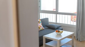 Een moderne woonkamer met een grijze bank, een witte salontafel met een fruitschaal, en een groot raam met gordijnen.