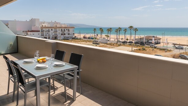 Un balcón con una mesa lista para dos personas, con vista a una playa con palmeras y un cielo despejado.