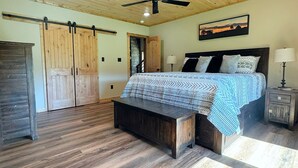 A bedroom with a bed, a bench, a ceiling fan, wooden flooring, and a painting on the wall.
